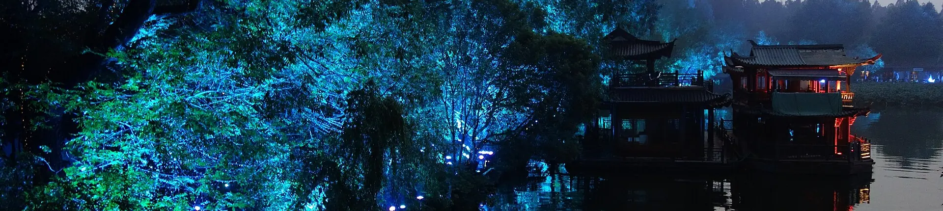 Blue-lit trees and a traditional pavilion reflected in a dark lake at night