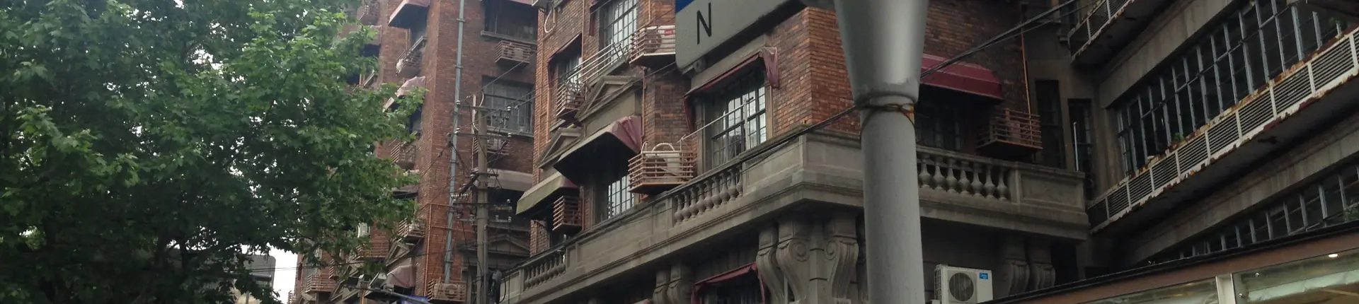 Balkone und graue Backsteinfassaden unter grünen Bäumen entlang der Wukang Road in Shanghai