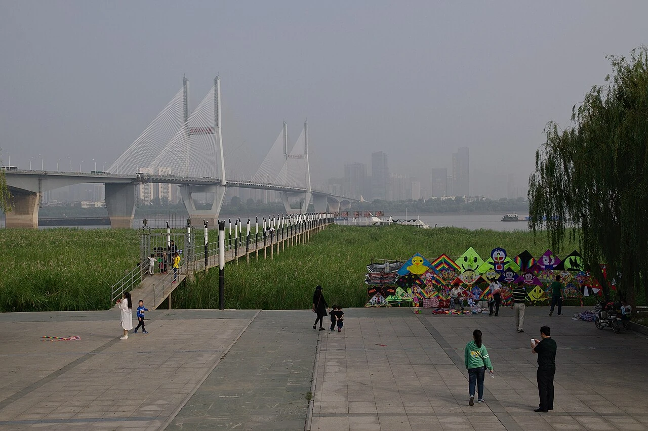 Visitors on a riverside promenade beside a cable-stayed bridge in hazy weather