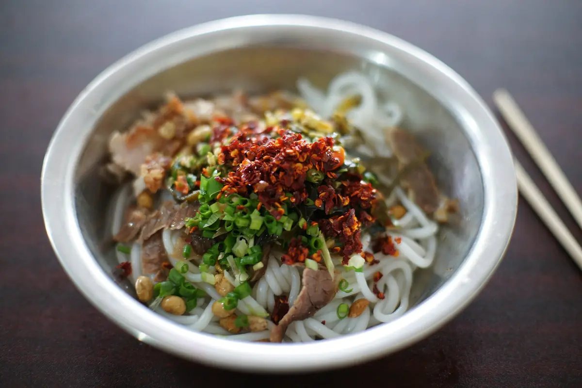 Rice noodles topped with chopped herbs, peanuts and red chili in a metal bowl