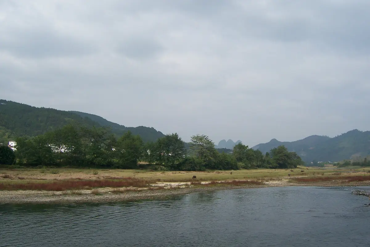 Shallow river beside a sandy bank and low wooded hills under gray clouds
