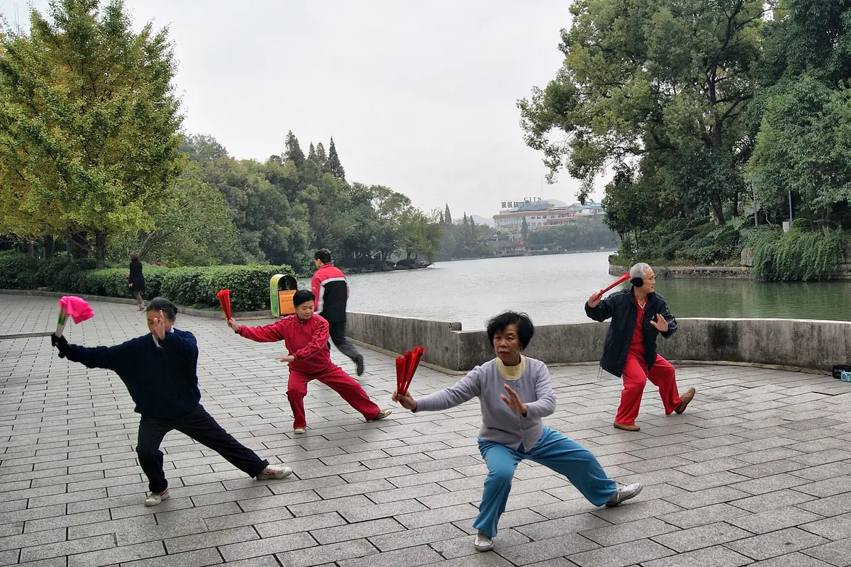 People practicing tai chi with swords on a paved lakeside terrace