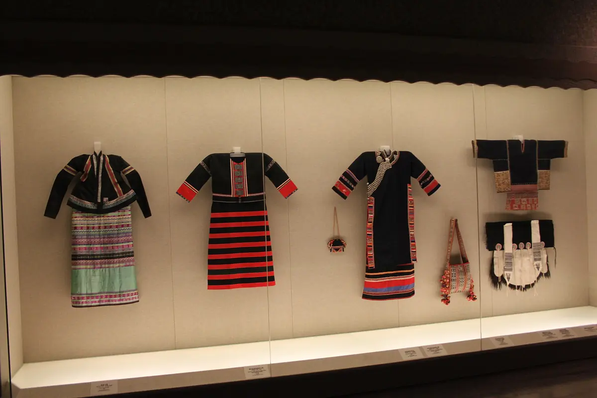 Trois robes traditionnelles à motifs et des accessoires textiles exposés derrière une vitre de musée