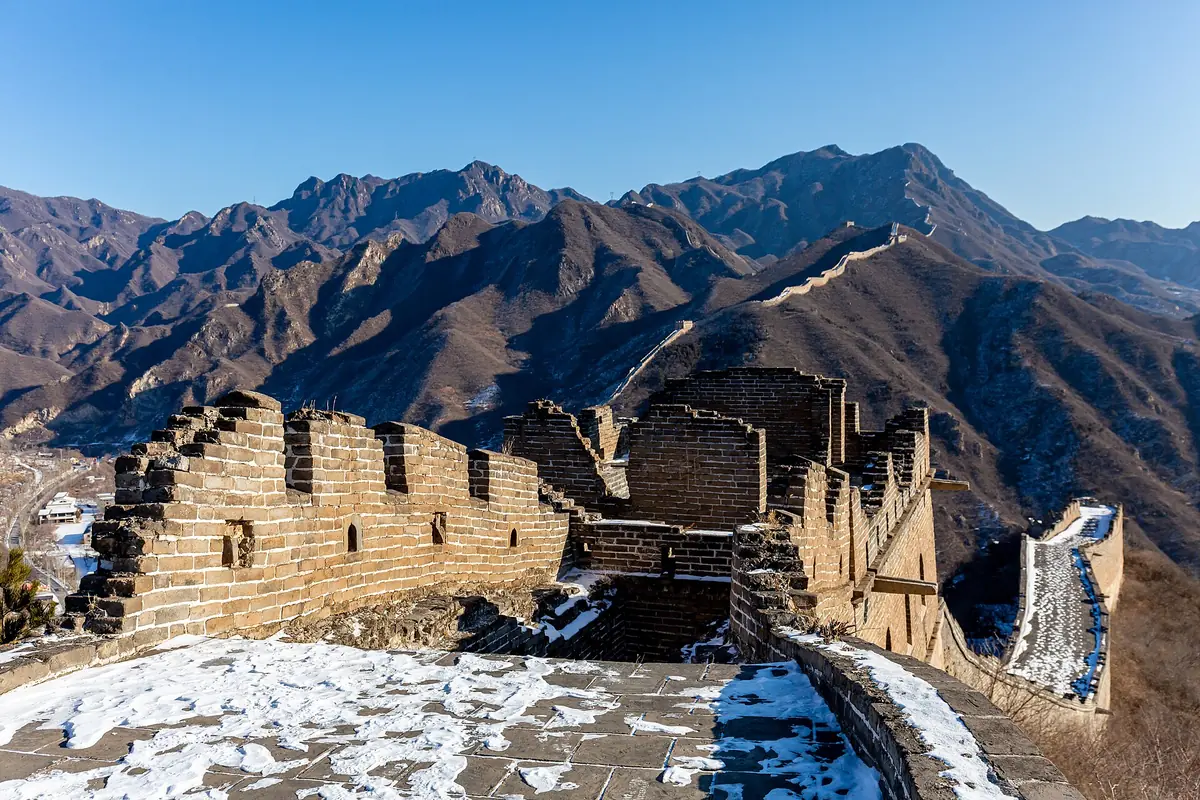 Snow-covered Great Wall ruins and a watchtower beneath rugged brown mountains