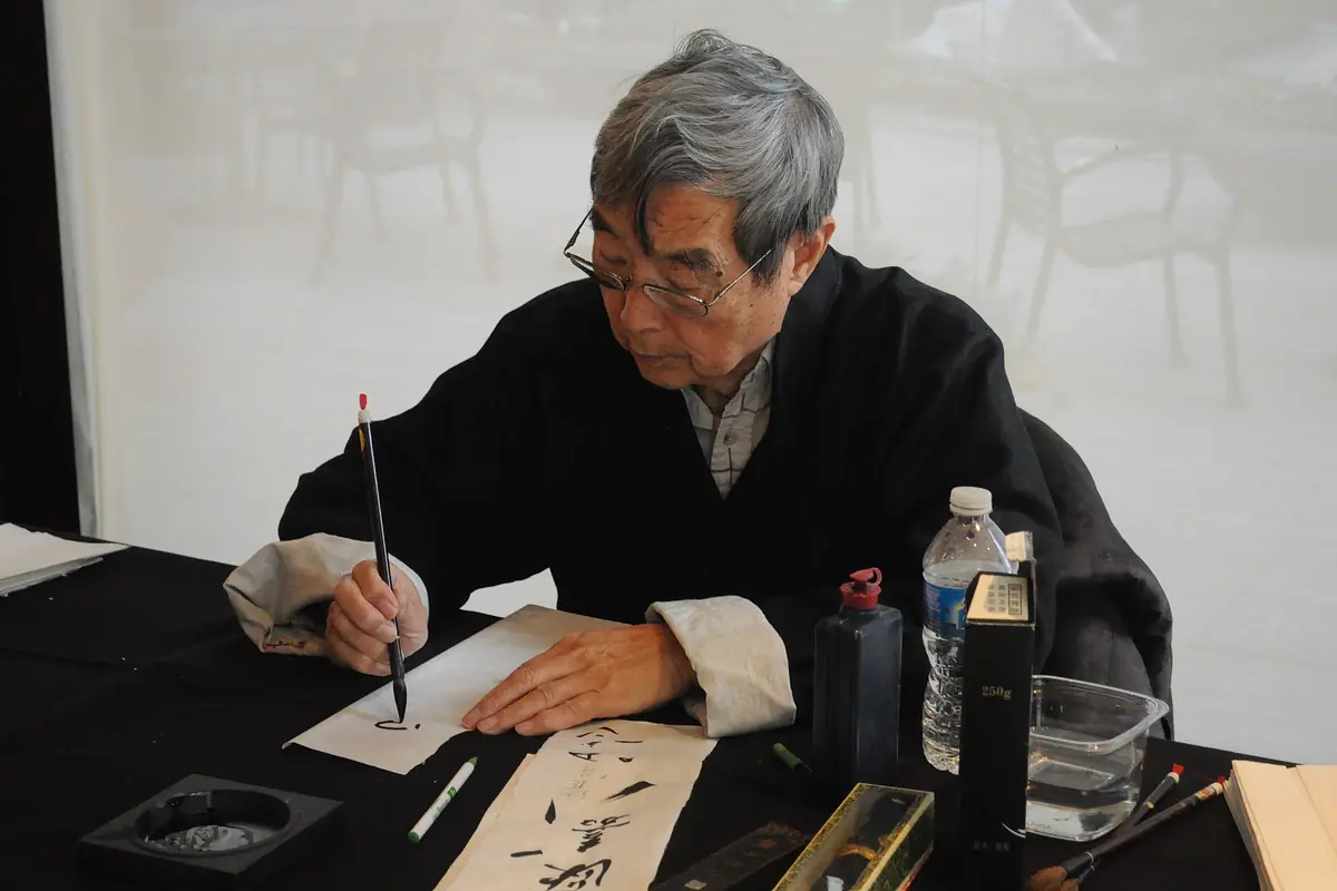 Older calligrapher writing Chinese characters with a brush at a table