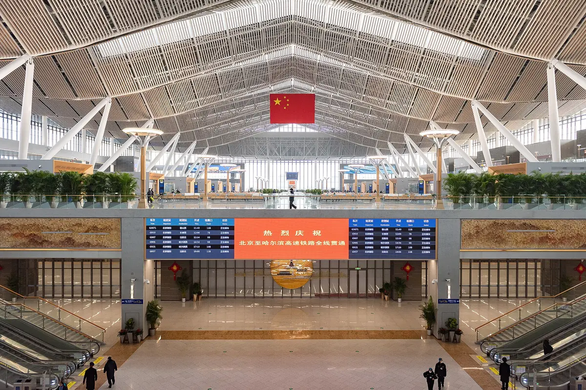 Salle d’attente ferroviaire avec des poutres de toit blanches, un drapeau chinois et des panneaux de départ