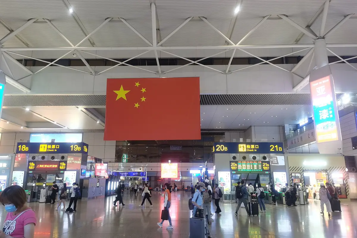 Passengers in a station concourse beneath a large Chinese flag and departure boards