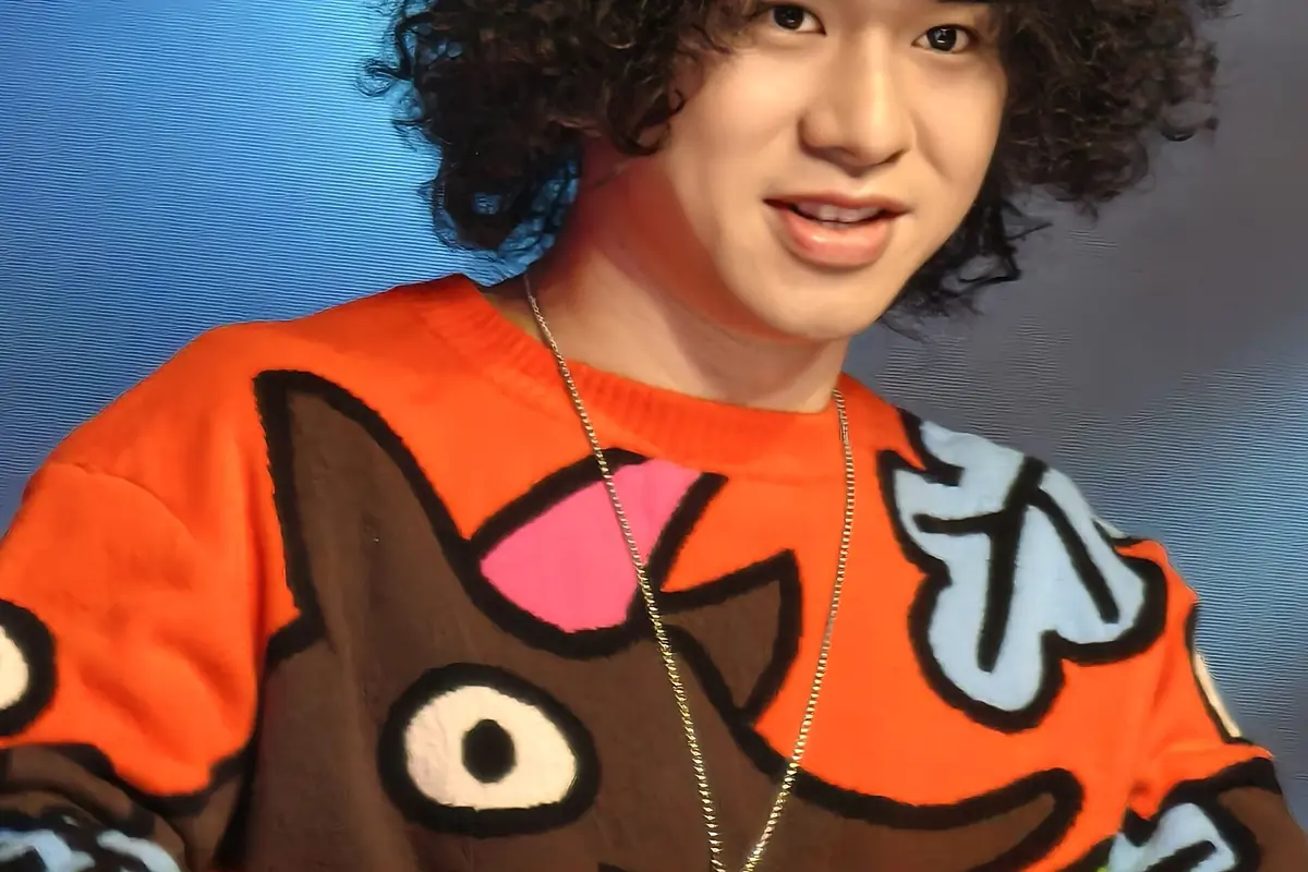 Curly-haired performer wearing an orange patterned sweater against a blue backdrop