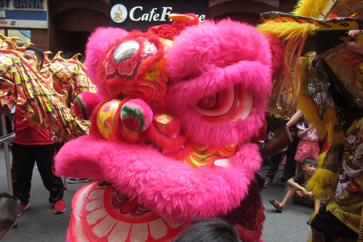 Bright pink lion-dance head with gold details facing a street crowd