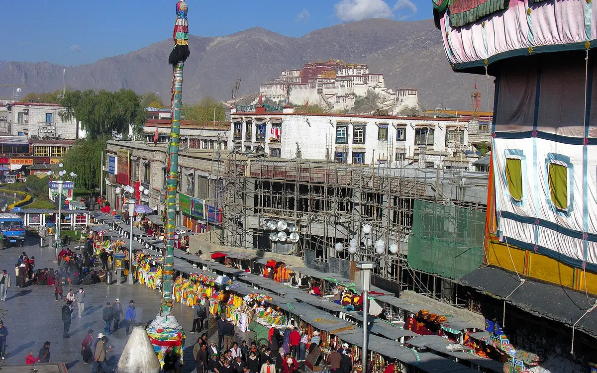 Busy Lhasa street with shops and crowds beneath a distant hilltop palace