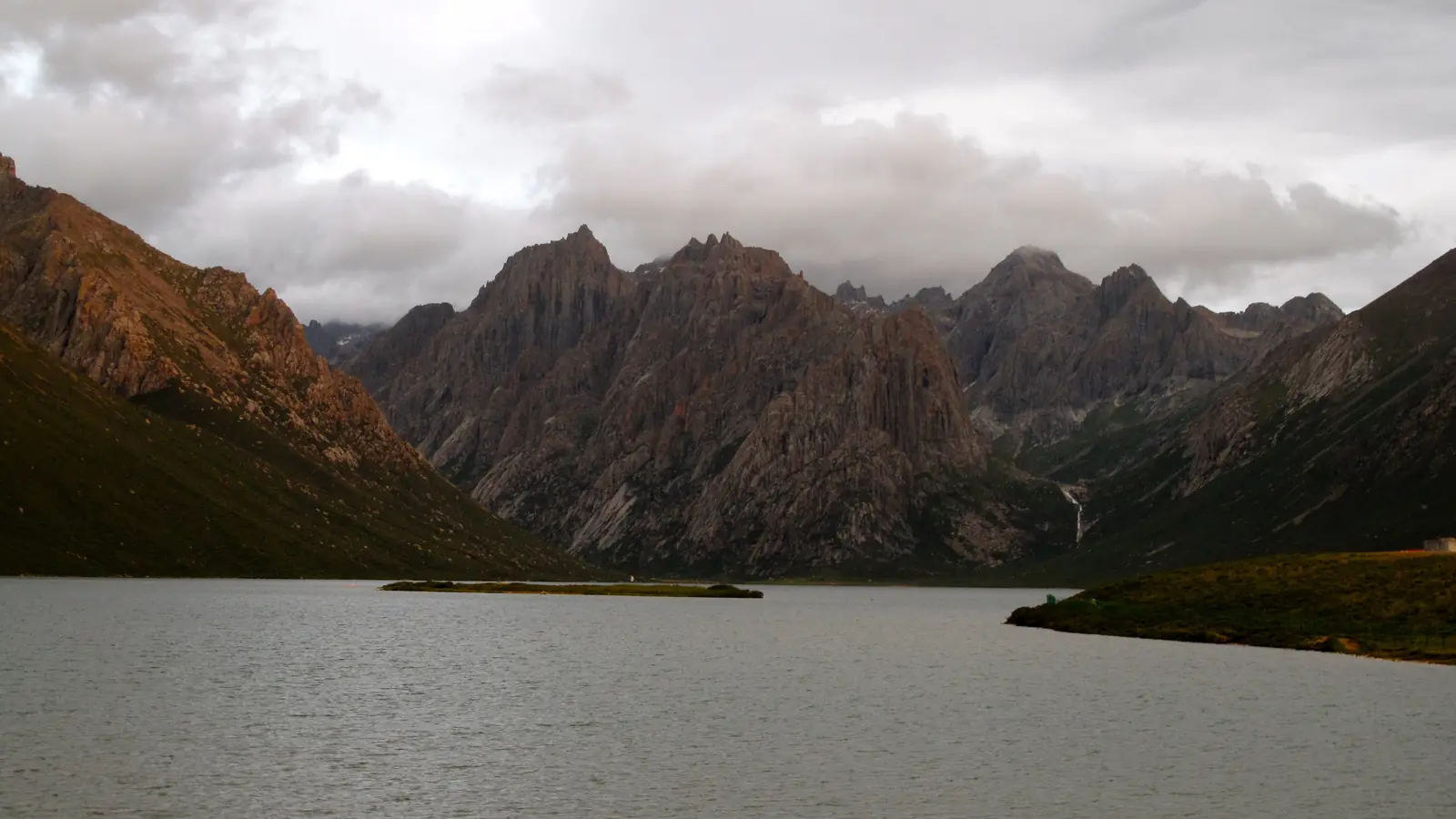 Jagged mountain ridges surrounding a high-altitude lake beneath clouds in Tibet
