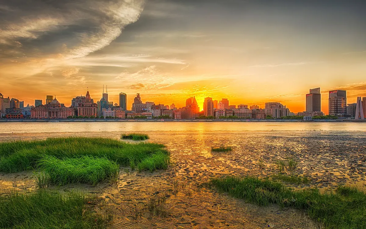 Sun setting behind a riverside city skyline beside a grassy shoreline
