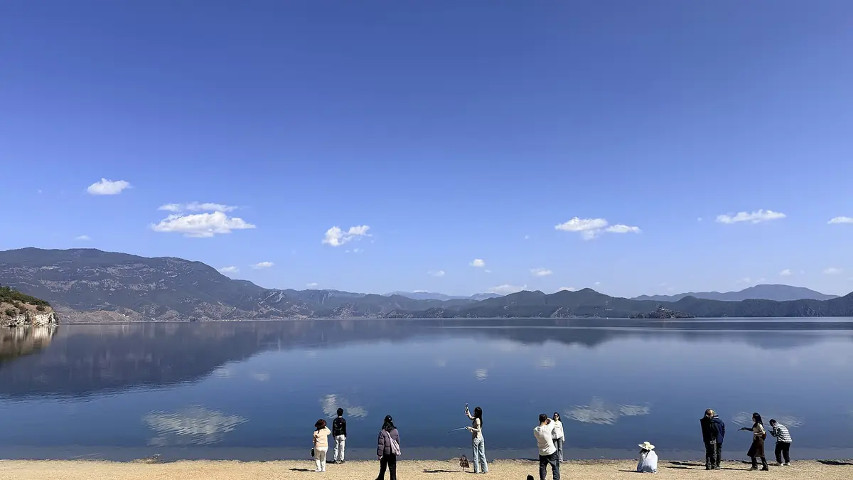 Visitors on a lakeshore beneath distant mountains and a clear blue sky